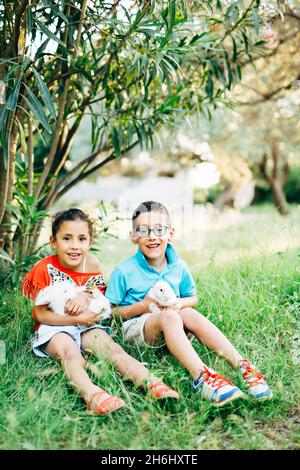 Istanbul, Türkei - 30.05.17: Lächelnder kleiner Junge und Mädchen, die im Garten unter einem grünen Busch auf dem Gras sitzen und weiße Kaninchen in den Armen halten Stockfoto