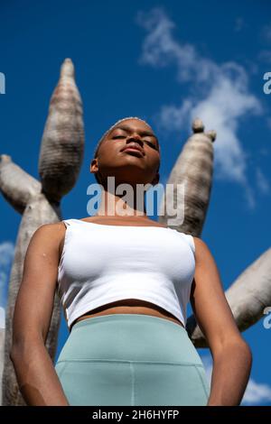Niedriger Winkel einer entspannten afroamerikanischen Frau, die mit geschlossenen Augen gegen den Drachenbaum steht und im Sommer sonniges Wetter im exotischen Garten genießt Stockfoto