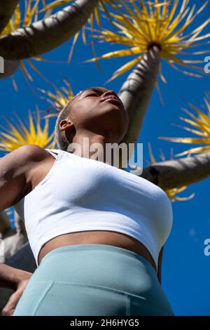 Von unten aus fitete afroamerikanische Sportlerin, die mit geschlossenen Augen im tropischen Garten mit Drachenbaum steht und den sonnigen Tag genießt Stockfoto