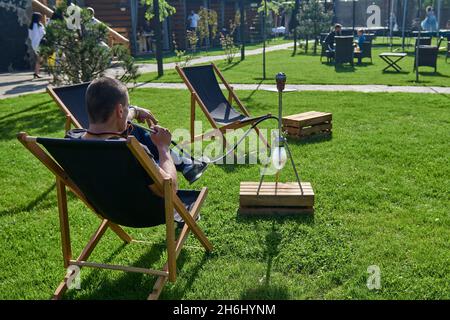 Mann, der an einem warmen sonnigen Tag Shesha, Shisha-Shisha in der Natur raucht. Östlicher Shisha Rauchen, aktive Erholung an der frischen Luft. Liegestühle auf geen Gras Stockfoto