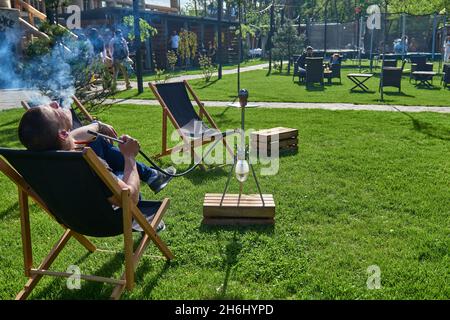 Mann, der an einem warmen sonnigen Tag Shesha, Shisha-Shisha in der Natur raucht. Östlicher Shisha Rauchen, aktive Erholung an der frischen Luft. Liegestühle auf geen Gras Stockfoto
