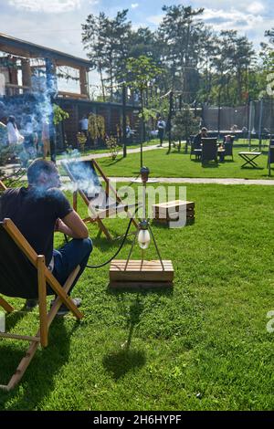 Mann, der an einem warmen sonnigen Tag Shesha, Shisha-Shisha in der Natur raucht. Östlicher Shisha Rauchen, aktive Erholung an der frischen Luft. Liegestühle auf geen Gras Stockfoto