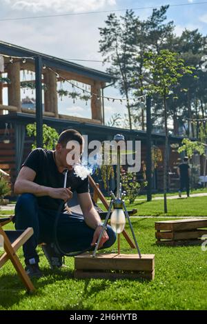 Mann, der an einem warmen sonnigen Tag Shesha, Shisha-Shisha in der Natur raucht. Östlicher Shisha Rauchen, aktive Erholung an der frischen Luft. Liegestühle auf geen Gras Stockfoto