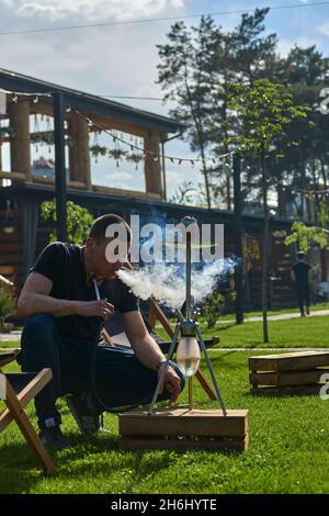 Mann, der an einem warmen sonnigen Tag Shesha, Shisha-Shisha in der Natur raucht. Östlicher Shisha Rauchen, aktive Erholung an der frischen Luft. Liegestühle auf geen Gras Stockfoto