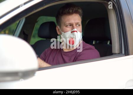 Ein Mann, der während eines Coronavirus-Ausbruchs in einem medizinischen Atemschutzgerät mit einem Auto fährt, ein Taxifahrer in einer Maske, schützt vor dem Virus. Fahrer im weißen Auto, Hand Stockfoto