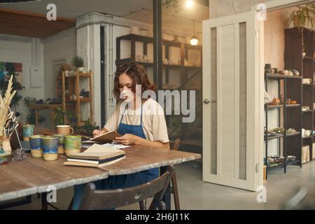 Junge Keramikerin in Arbeitskleidung, die Notizen macht, während sie in ihrer Werkstatt am Tisch vor dem Laptop sitzt Stockfoto