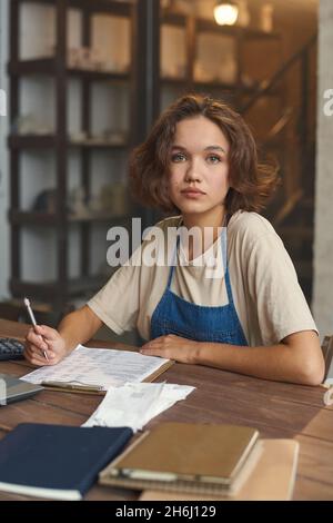 Junge hübsche Keramikerin, die in ihrer Werkstatt mit Dokumenten am Tisch arbeitet und die Kamera anschaut Stockfoto