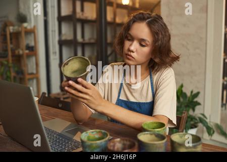 Junge kreative Kunsthandwerkerin in Arbeitskleidung, die selbstgemachten Tonbecher hält und ihn an ihrem Arbeitsplatz ansieht Stockfoto