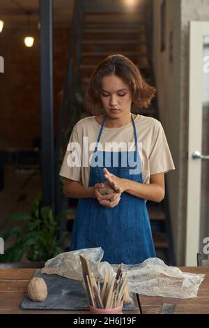 Junge Handwerkerin in Arbeitskleidung knetet ein Stück Ton zwischen den Händen und bereitet es für die weitere Verarbeitung vor Stockfoto