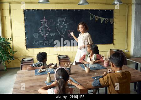 Junge selbstbewusste Lehrerin erklärt Schülern, die vor der Tafel an einem großen Tisch sitzen, Skizzen der Töpferei Stockfoto