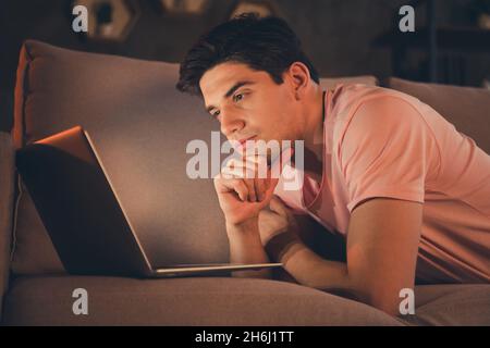 Profilansicht eines attraktiv gesinnten, fokussierten Mannes, der auf einem Sofa mit einem Laptop liegt und den Finanzbericht zuhause liest Stockfoto