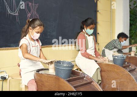 Reihe von Schülern, die an einer Tafel sitzen und Tontöpfe vor Töpferscheiben in einem großen Klassenzimmer oder einer Werkstatt modellieren Stockfoto