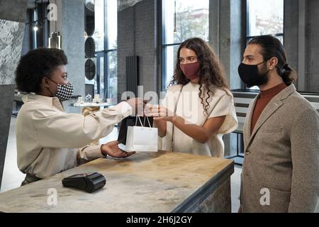 Junge Frau in Schutzmaske nimmt Paperbags mit Kauf durch afrikanische weibliche Shop-Assistentin über den Zähler im Juweliergeschäft Stockfoto