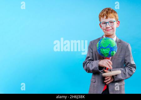 Schuljunge trägt Anzug und Krawatte mit einer Brille, die sein wissenschaftliches Projekt „Globe of Earth“ auf blauem Hintergrund hält, ohne Platz für Text Stockfoto