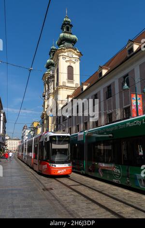 Linz, Österreich - 26. September 2021: Altstadt von Linz mit bunten Straßenbahnen, die vor einer historischen Kirche vorbeifahren Stockfoto