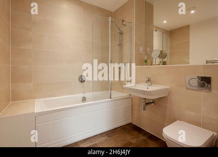 Cambridge, England - Aug 6 2019: Moderne, teilweise geflieste Dusche/Bad mit wandhängendem Keramikwaschbecken, Badewanne und großem Spiegel Stockfoto
