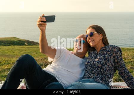 Gealtertes Paar, das auf der Wiese liegt, Selfies macht und den Sonnenuntergang an der Küste genießt Stockfoto