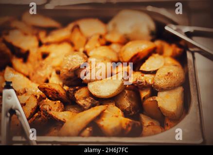 In der Fritteuse befinden sich viele leckere, appetitlich gebratene rustikale Kartoffeln, daneben gibt es Zangen. Kalorienreiche Lebensmittel und Fast Food. Junk Food. Stockfoto