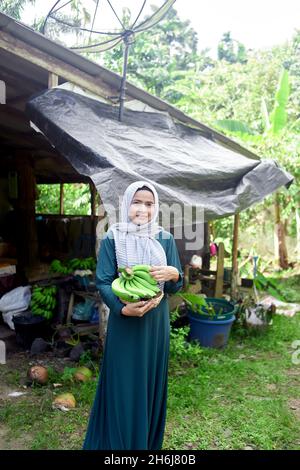 Muslimische Frau im Hijab, die im Heimatbereich steht und rohe Banane hält Stockfoto