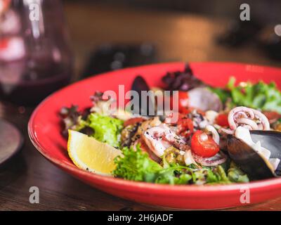 Frischer mediterraner Seafood-Salat mit Olivenöl, Zitrone, Tomaten und Salat in roter Platte auf dem Tisch im Restaurant Stockfoto