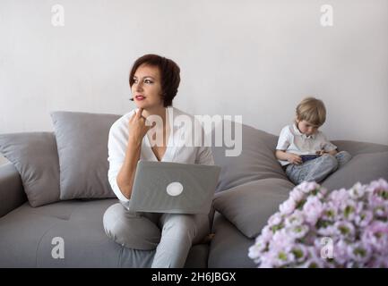 Mutter und Sohn mit digitalen Gadgets im Wohnzimmer. Eine Frau arbeitet zu Hause auf einem Laptop, neben einem Kind spielt am Telefon. Das Konzept der Moderne Stockfoto