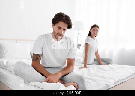 Aufgeregt beleidigt kaukasischen jungen Mann ignoriert Frau im Bett im weißen Schlafzimmer Innenraum sitzt, entschuldigt sich die Dame Stockfoto