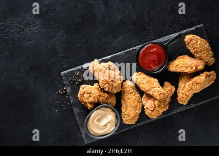 Knusprig gebratene Hähnchenflügel mit Saucen auf schwarzem Schieferbrett, Draufsicht, Kopierfläche. Junk Food Stockfoto