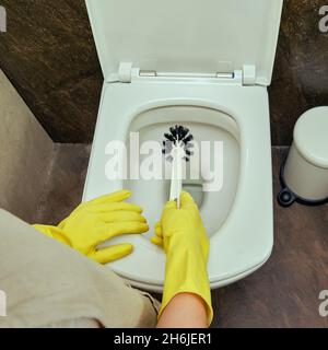 Eine Frau putzt das Badezimmer und wischt die Toilettenschüssel mit einer Bürste ab. Desinfektion mit gelben Gummihandschuhen im Waschraum Stockfoto