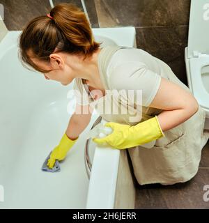 Die Frau putzt das Badezimmer und wäscht die Badewanne. Desinfektion mit gelben Gummihandschuhen im Waschraum Stockfoto
