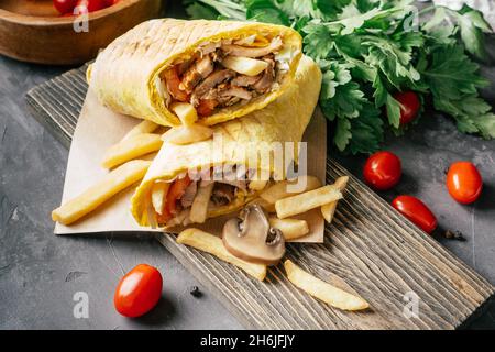 Rustikale Rolle Shawarma mit pommes frites, Champignons, Huhn und Gemüse in Pita-Brot auf dunklem Holzhintergrund mit Kirschtomaten und grünem pa Stockfoto