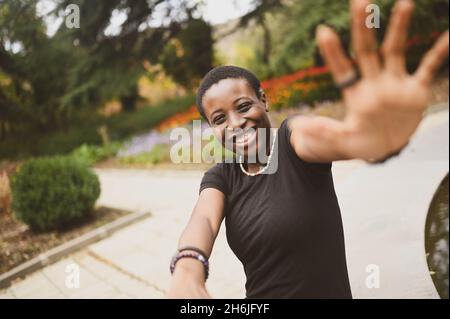 Nahaufnahme Porträt von attraktiven glücklich lächelnd junge natürliche Schönheit kurzhaarige afrikanische Frau trägt schwarzes T-Shirt lachend Strecke die Hände aus schließen von der Kamera in Natur Sommerpark Stockfoto