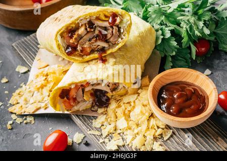 Knusprige Shawarma-Rolle mit Chips, Grillsauce, Huhn und Gemüse in Pitabrot auf dunklem Holzhintergrund mit Kirschtomaten und grünen Pars Stockfoto