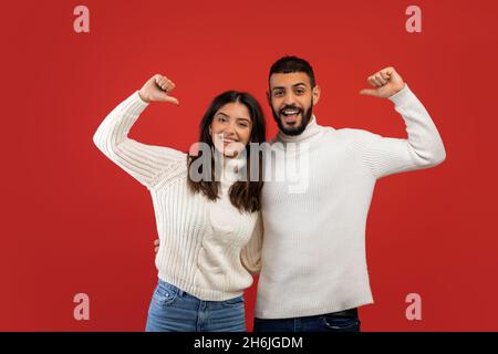Glückliche junge arabische Ehegatten in weißen Pullovern posieren und zeigen Daumen auf sich selbst, roter Hintergrund Stockfoto