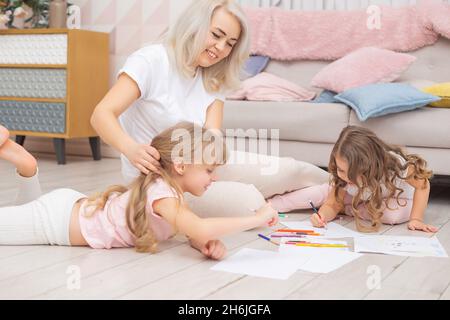 Die 30-jährige kaukasische Mutter hilft ihren niedlichen kleinen Töchtern beim Zusammenziehen, die im Wohnzimmer auf dem Boden sitzen. Glückliches Familienkonzept. Stockfoto