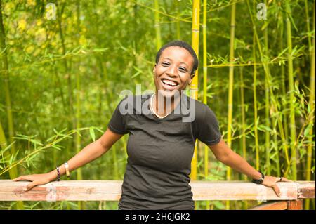 Attraktive glücklich lächelnde junge natürliche Schönheit kurzhaarige afrikanische Schwarze Frau lachend lehnt sich an ein Holzgeländer posiert vor dem Hintergrund der Bambushain im Natur Sommerpark Stockfoto