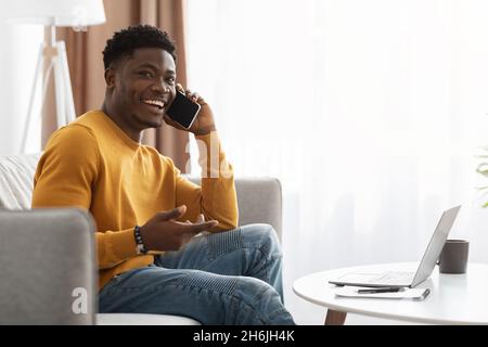 Fröhlicher schwarzer Kerl, der von zu Hause aus arbeitet und Telefongespräche führt Stockfoto