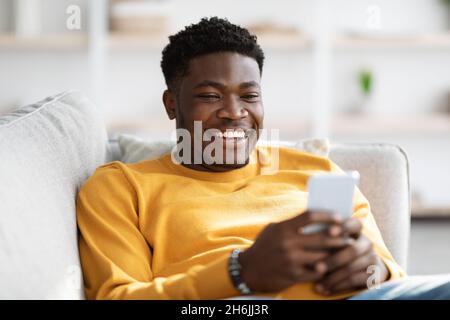 Lächelnder afroamerikanischer Mann, der mit dem Handy auf der Couch liegt Stockfoto