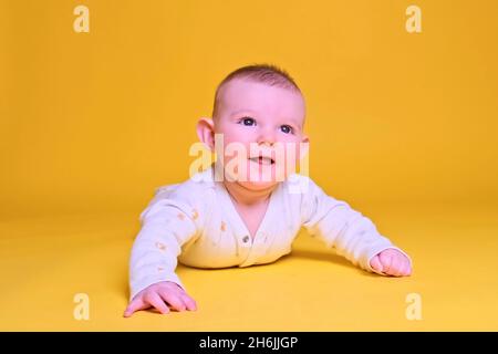 Fröhlicher kleiner Junge, der lächelnd auf seinem Bauch liegt, gelber Hintergrund im Studio. Vier Monate altes Kind aus nächster Nähe Stockfoto