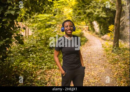 Attraktive glücklich lächelnde junge natürliche Schönheit kurzhaarige afrikanische schwarze Frau mit blauen Kopfhörern in schwarzem T-Shirt Musik hören und tanzen in Natur Sommerpark Stockfoto