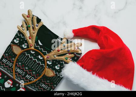 Frohe Weihnachten. Draufsicht auf weihnachts-Pullover mit hellen weihnachts-Spielzeug-Hirschgeweih und Weihnachtsmann-Hut auf einem Marmortisch Stockfoto