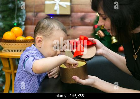 Mutter und Sohn zerlegen eine Schachtel mit Neujahrsgeschenken auf dem Hintergrund des geschmückten Weihnachtsbaums. Das Konzept des neuen Jahres. Stockfoto