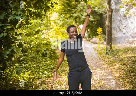 Attraktive glücklich lächelnde junge natürliche Schönheit kurzhaarige afrikanische Frau trägt total schwarz Wandern und Tanzen in Natur grünen Sommerpark. Diversity-Konzept Stockfoto