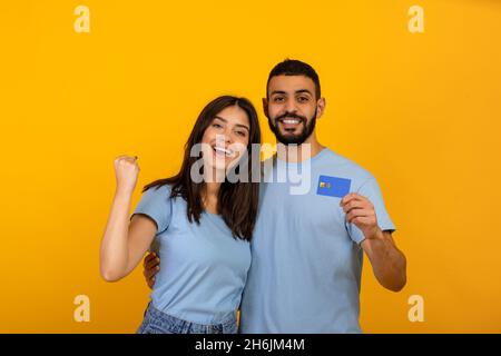 Überfröhliche arabische Paare mit Kreditkarte, die eine „JA“-Geste machen und kontaktloses Einkaufen auf gelbem Hintergrund fördern Stockfoto