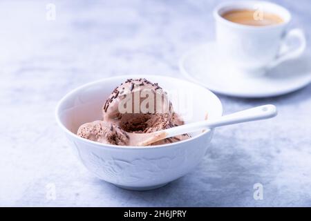 Schokoladeneis mit Schokolade in einer weißen Schüssel auf einem hellen Hintergrund streuen. Drei Kugeln hausgemachtes Eis und eine Tasse Kaffee. Nahaufnahme. Stockfoto