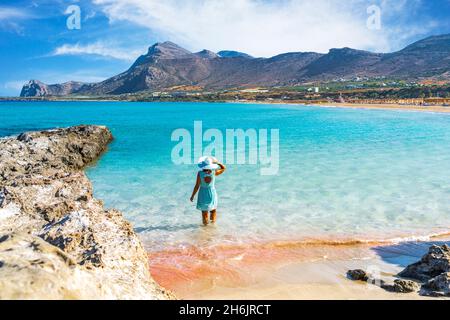 Frau in Kleid, die im türkisklaren Meer mit Blick auf den rosa Sandstrand von Falassarna, Kreta, griechische Inseln, Griechenland, Europa, steht Stockfoto
