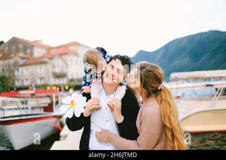 Lächelnder Vater trägt seine Tochter auf seinen Schultern. Mutter und Tochter küssen Vater von beiden Seiten. Mädchen hält eine Spielzeugblume in den Händen Stockfoto
