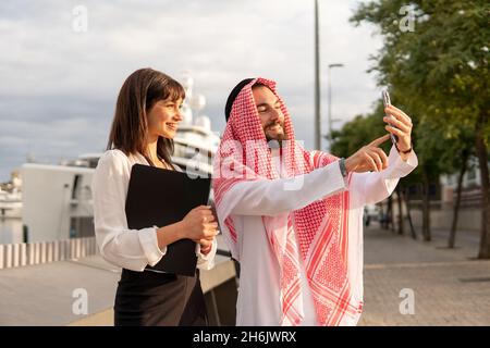 Lächelnder arabischer Geschäftsmann in traditioneller saudischer Kleidung, der im Freien Selfie mit einer jungen attraktiven Kollegin macht, glückliche und vielfältige Geschäftspartner, die im Hafen Fotos mit dem Mobiltelefon machen Stockfoto