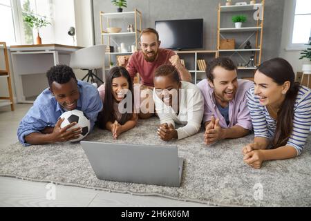 Fröhliche Freunde, eine vielfältige Gruppe, die Fußballspiele auf einem Laptop auf dem Boden beobachtet Stockfoto