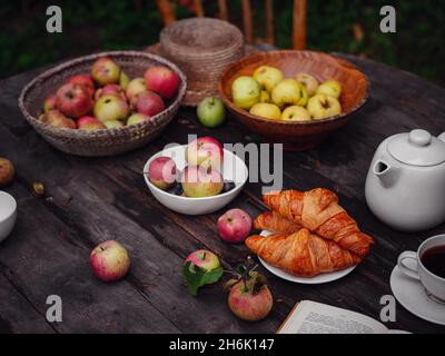 Schöne Herbst Stillleben in Apfelgarten, alten Holztisch mit Becher und Teekannen, Äpfel und Pflaumen, Croissants und offenes Buch, Idee und Konzept der h Stockfoto