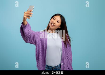 Nette asiatische Dame, die Selfie auf dem Smartphone macht, mit verspielter Stimmung und lächelnd auf die Webcam, blauer Hintergrund Stockfoto